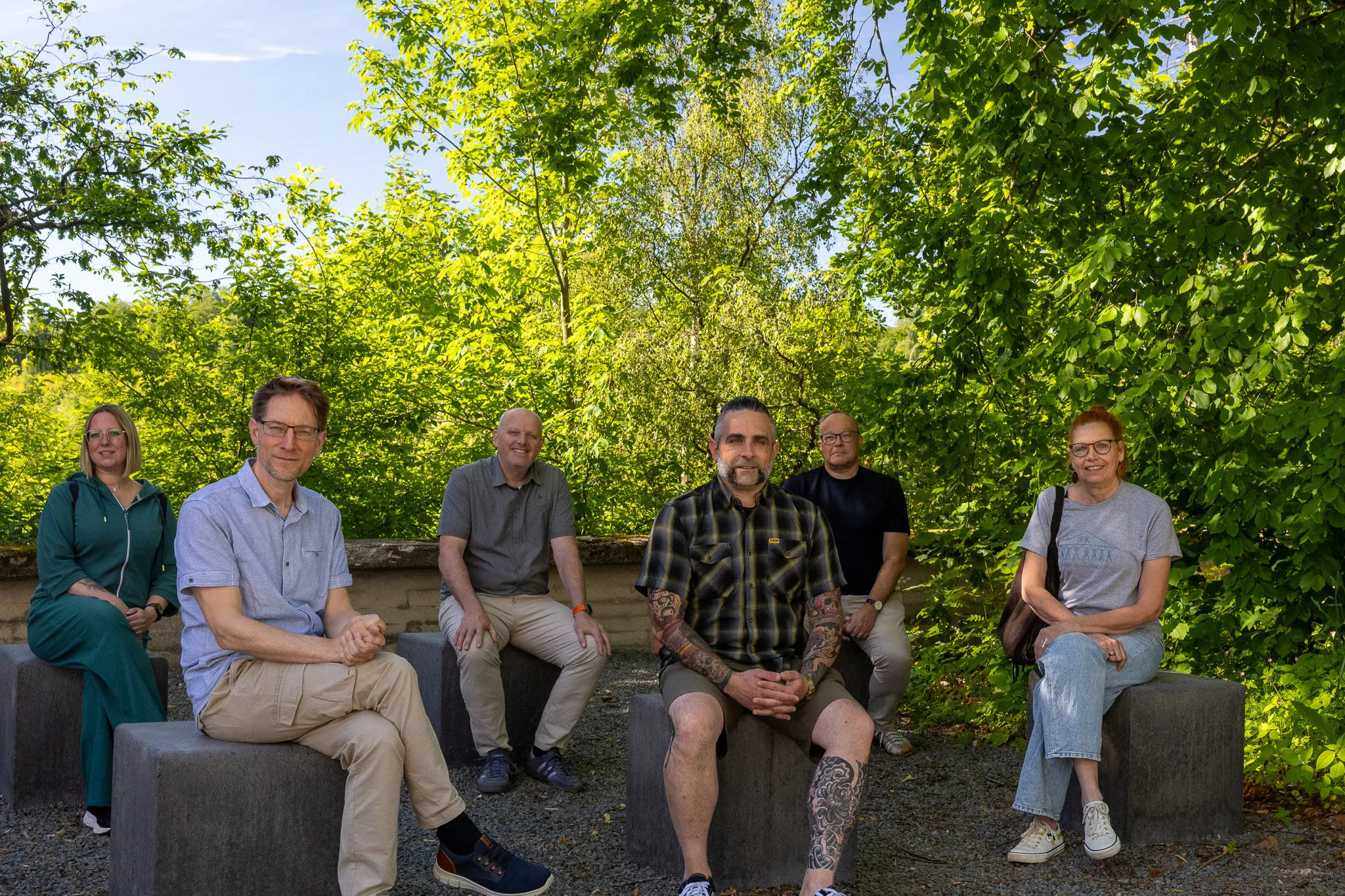 Sechs Personen sitzen im Freien auf grauen Betonblöcken vor üppigem Grün. Sie spenden 2500 Euro für Obdachlosenhilfe.