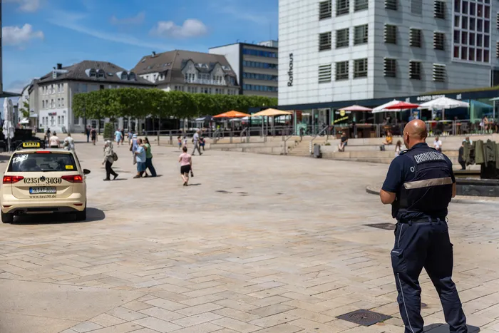 Ein Polizist mit der Aufschrift „Ordnungsamt“ steht mit dem Rücken zur Kamera auf einem Platz und blickt in die Ferne. Im Hintergrund sind Gebäude, Fußgänger und ein Taxi zu sehen.
