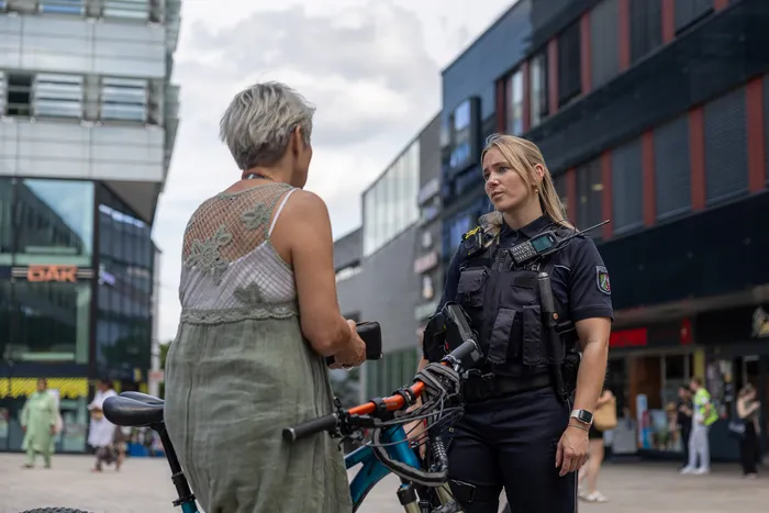 Polizistin spricht eine ältere Radfahrerin in einer Fußgängerzone an. Im Hintergrund sind moderne Gebäude und Passanten zu sehen.