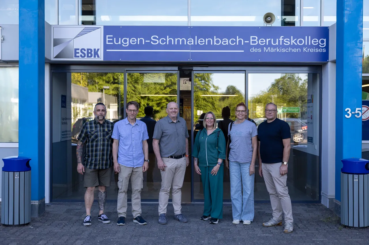 Fünf Personen stehen vor dem Eingang des Eugen-Schmalenbach-Berufskollegs, darunter ein Mann mit Bart und eine Frau mit roten Haaren. Im Hintergrund ist das Logo des ESBK zu sehen.