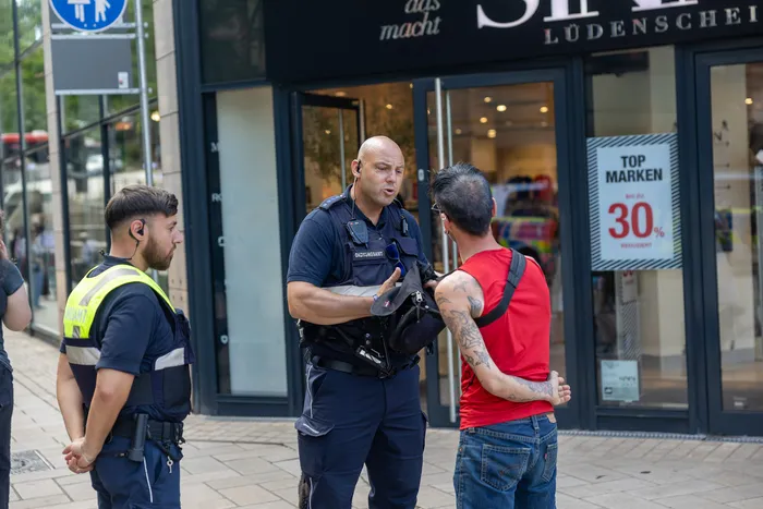 Polizisten kontrollieren einen jungen Mann mit Tattoos in der Fußgängerzone von Lüdenscheid. Im Hintergrund ist ein Schaufenster mit einem Werbeplakat für reduzierte Markenartikel zu sehen.