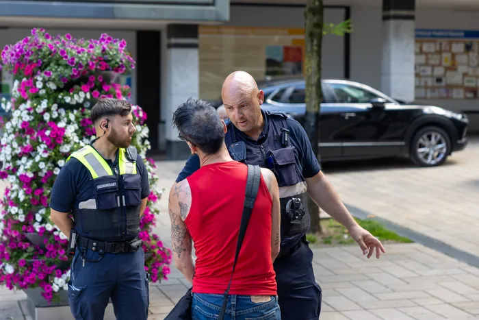 Zwei Polizisten sprechen einen Mann in rotem Tanktop und Jeans an. Im Hintergrund sind ein Auto und blühende Pflanzen zu sehen.