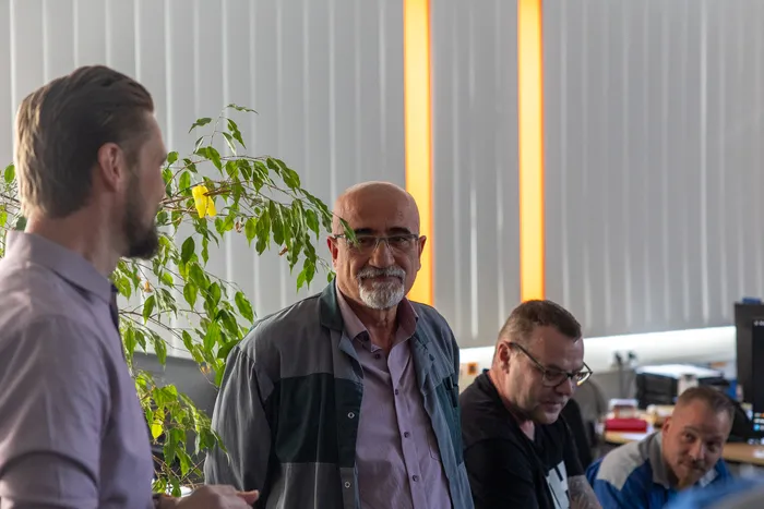 Ein Mann mit grauen Haaren und Brille blickt lächelnd in die Kamera, während ein Kollege im Hintergrund mit ihm spricht. Im Hintergrund sind weitere Personen an Arbeitsplätzen zu sehen.