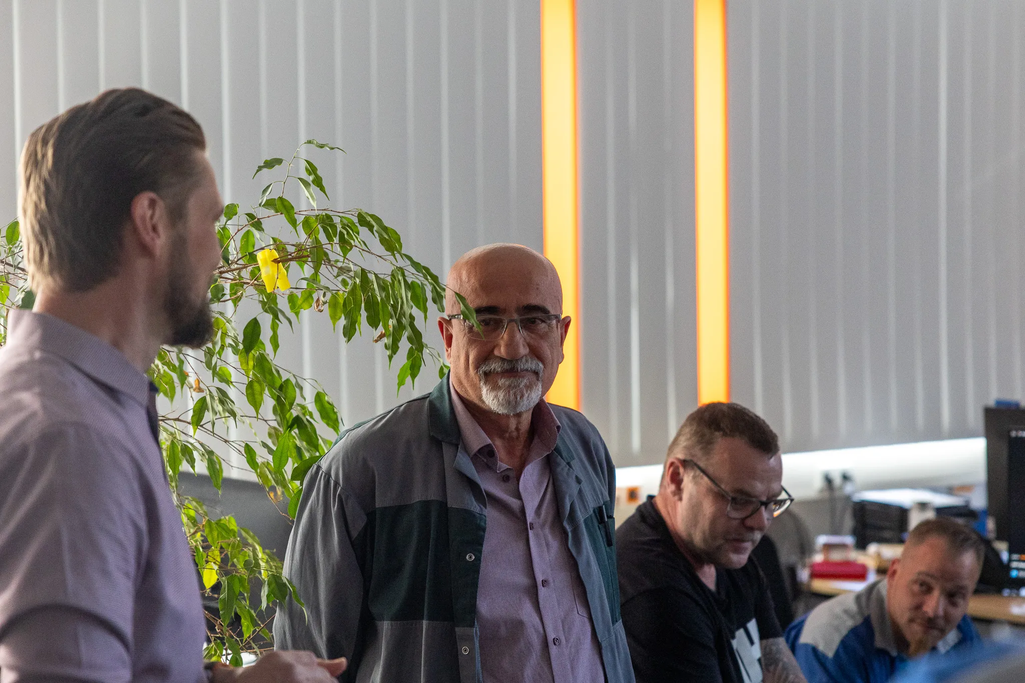 Ein Mann mit grauen Haaren und Brille blickt lächelnd in die Kamera, während ein Kollege im Hintergrund mit ihm spricht. Im Hintergrund sind weitere Personen an Arbeitsplätzen zu sehen.