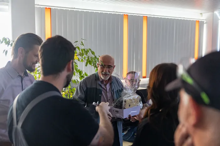Ein älterer Mann mit Bart nimmt eine Geschenkbox von Kollegen entgegen, umgeben von weiteren Mitarbeitenden in einem hellen Raum.