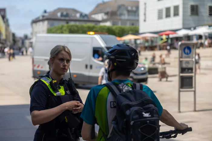 Polizistin spricht einen Radfahrer im Stadtzentrum an. Im Hintergrund sind weitere Fußgänger und Fahrzeuge zu sehen.