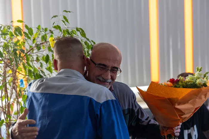 Zwei Männer umarmen sich herzlich, einer hält einen bunten Blumenstrauß. Im Hintergrund sind Pflanzen und orangefarbene Lichtstreifen zu sehen.
