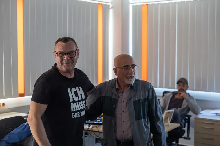Zwei Männer im Vordergrund, einer trägt ein T-Shirt mit der Aufschrift „Ich muss gar nix“, der andere eine Jacke. Im Hintergrund sind weitere Personen in einem Büro zu sehen.