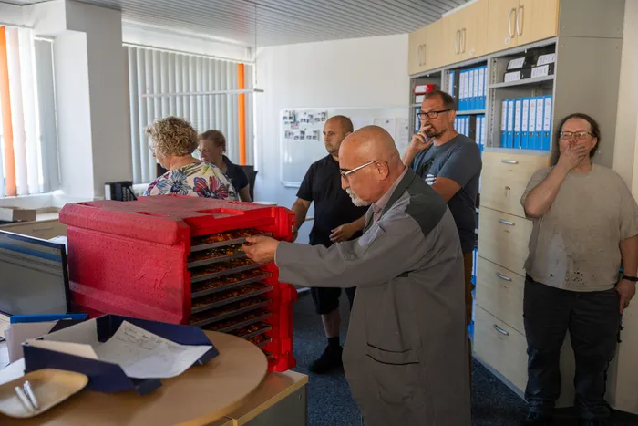 Ein älterer Mann in Arbeitskleidung öffnet eine rote Trocknungsanlage mit mehreren Ebenen, umgeben von Kollegen in einem Büro.