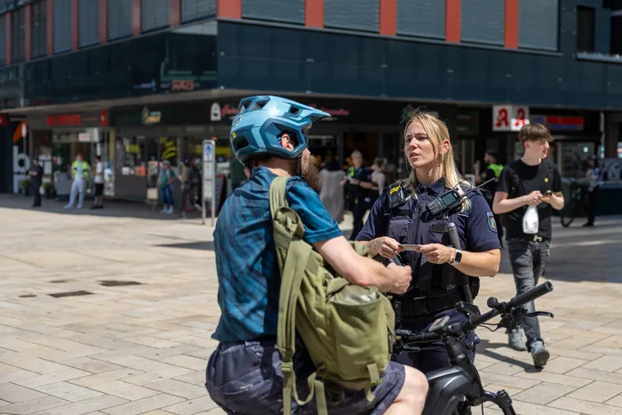 Polizistin kontrolliert einen Radfahrer mit Helm und Rucksack in einer Fußgängerzone. Im Hintergrund sind Passanten und Geschäfte zu sehen.