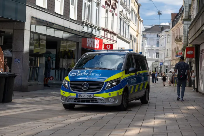 Ein blauer Polizei-Transporter der Marke Mercedes steht auf einem Pflastersteinweg in einer Fußgängerzone. Im Hintergrund sind Geschäfte und Passanten zu sehen.
