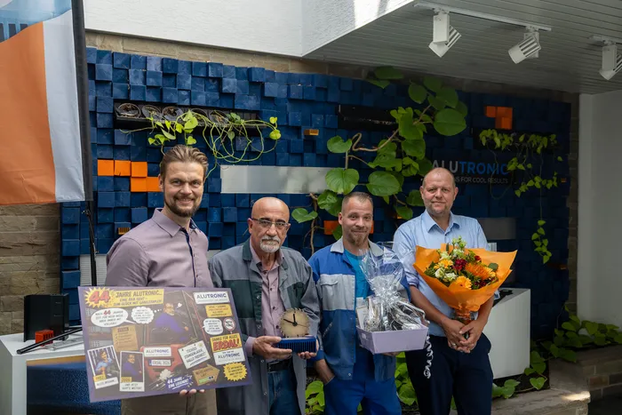 Drei Männer und ein Junge stehen vor einer Wand mit Pflanzen und Werbematerialien. Der Mann in der Mitte hält eine Auszeichnung, der Mann rechts hält einen Blumenstrauß.