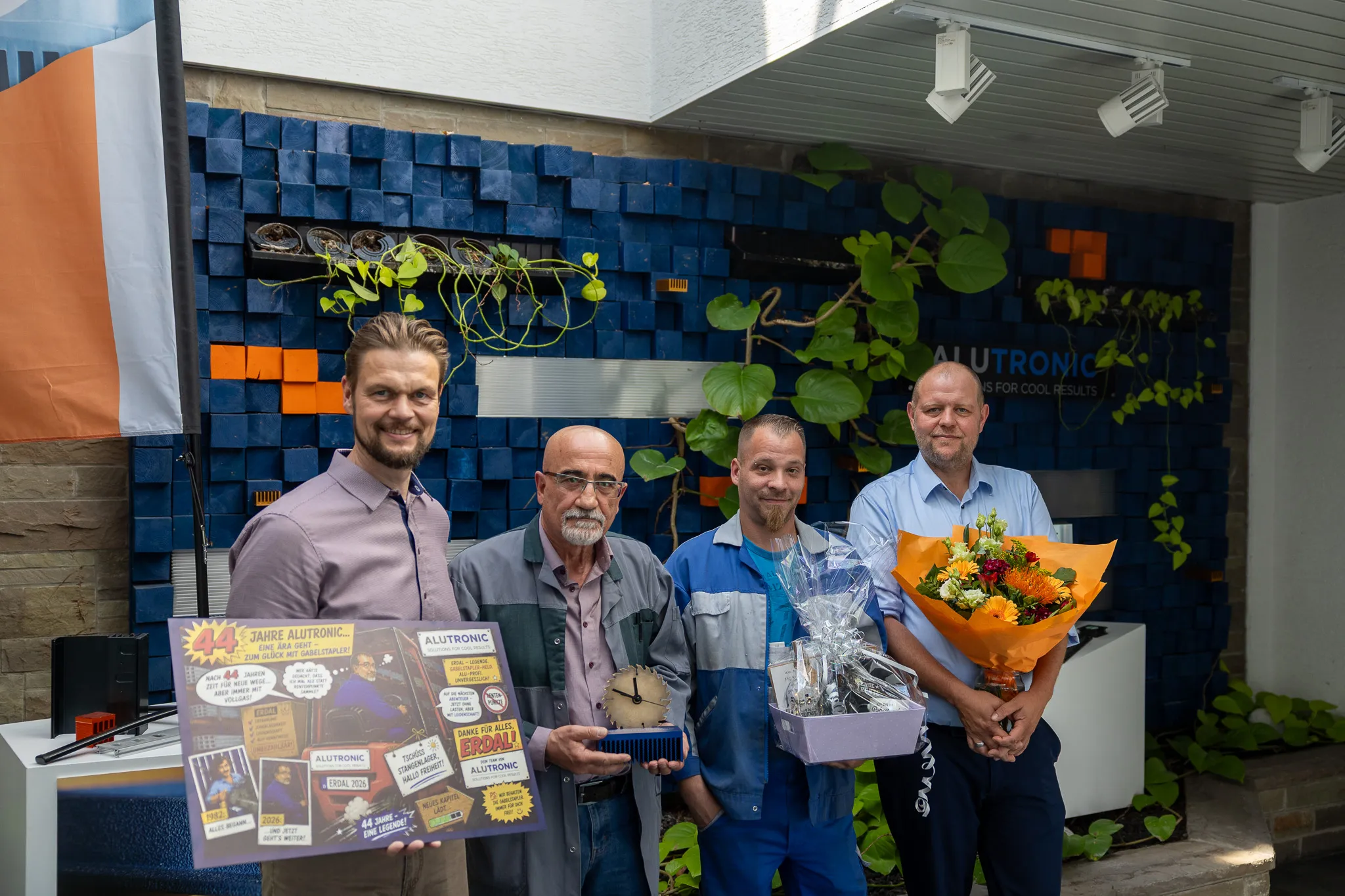 Drei Männer und ein Junge stehen vor einer Wand mit Pflanzen und Werbematerialien. Der Mann in der Mitte hält eine Auszeichnung, der Mann rechts hält einen Blumenstrauß.