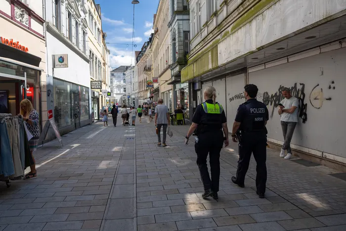 Zwei Polizisten in Uniform gehen auf einer belebten Fußgängerzone in Lüdenscheid entlang. Im Hintergrund sind Geschäfte und Passanten zu sehen.