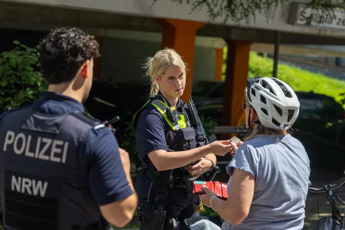 Polizisten kontrollieren eine Radfahrerin auf einem Bürgersteig. Eine Beamtin nimmt einen Ausweis entgegen, während ihr Kollege im Hintergrund steht.