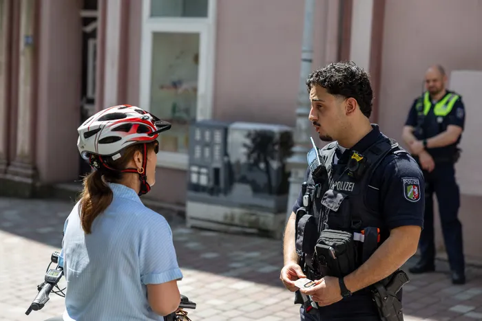 Ein Polizist spricht eine Radfahrerin in der Fußgängerzone an. Er hält ein Tablet in der Hand, während die Radfahrerin einen Helm trägt und ihr Fahrrad im Hintergrund steht. Ein weiterer Polizist ist im Hintergrund zu sehen.