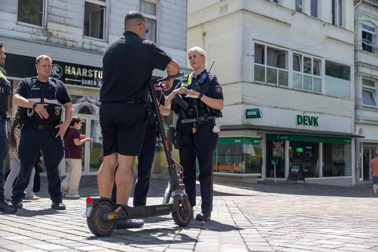 Polizisten kontrollieren einen Mann mit einem E-Scooter in einer Fußgängerzone. Im Hintergrund sind Geschäfte und weitere Personen zu sehen.