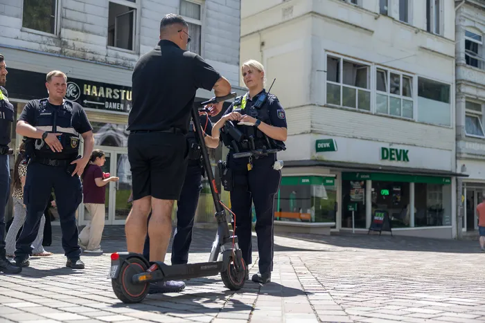 Polizisten kontrollieren einen Mann mit einem E-Scooter in einer Fußgängerzone. Im Hintergrund sind Geschäfte und weitere Personen zu sehen.