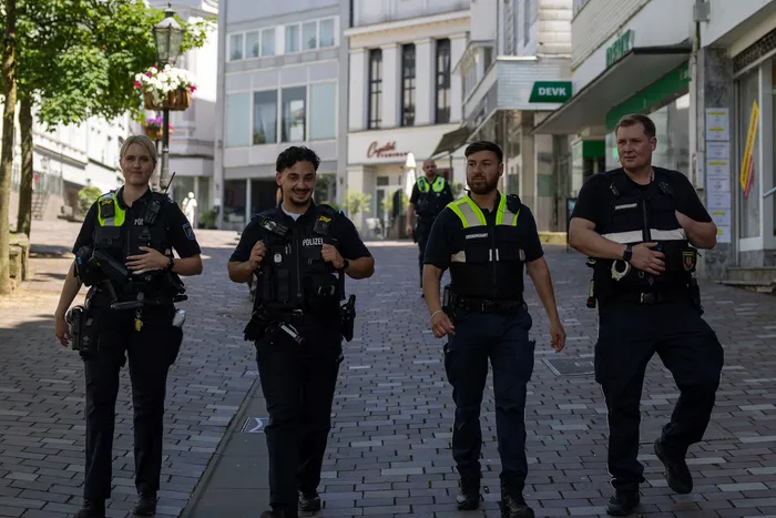Vier Polizisten in Uniform gehen auf eine Straße zu. Zwei von ihnen tragen Warnwesten. Im Hintergrund sind Geschäfte und Gebäude zu sehen.