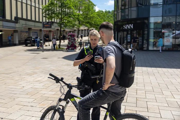 Eine Polizistin in Uniform kontrolliert auf einer Fußgängerzone einen Mann, der neben seinem Fahrrad steht und ebenfalls auf sein Smartphone schaut. Im Hintergrund sind Geschäfte und Passanten zu sehen.
