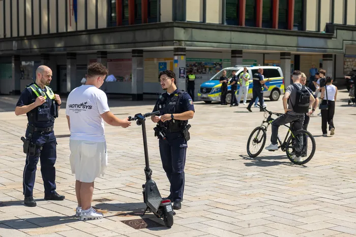 Polizisten kontrollieren einen jungen Mann auf einem E-Scooter in einer Fußgängerzone. Im Hintergrund sind weitere Beamte und Passanten zu sehen.