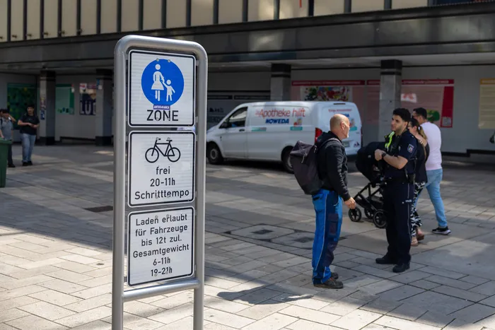 Schild mit Piktogrammen für Fußgängerzone, Fahrradverkehr und Lademöglichkeiten steht auf einem Platz. Im Hintergrund sind Menschen und Fahrzeuge zu sehen, sowie zwei Polizisten im Gespräch.