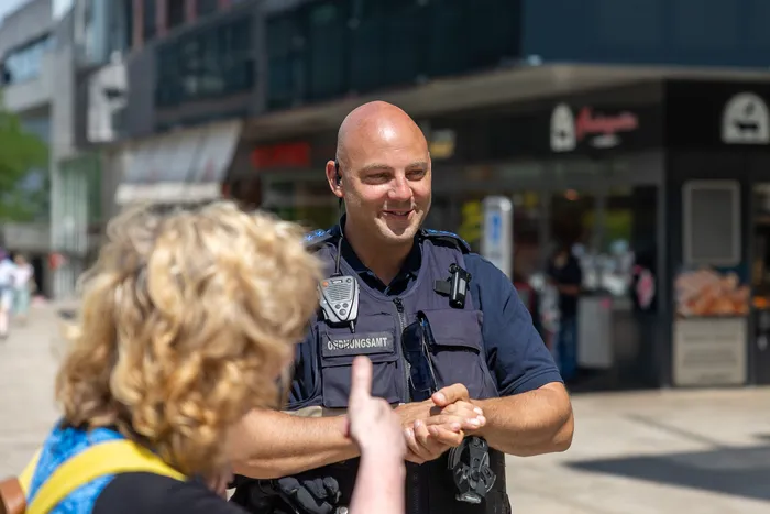 Ein Polizist im Dienst lächelt und nimmt die Hände einer Person entgegen. Im Hintergrund sind Gebäude und Passanten in einer Fußgängerzone zu sehen.