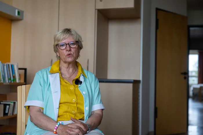 Eine Frau mit kurzem blondem Haar und Brille sitzt vor einem hellen Hintergrund. Sie trägt ein gelbes Oberteil mit einem hellgrünen Jackett und hält ihre Hände vor sich zusammen.