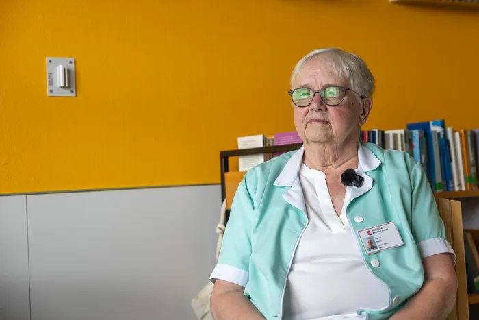 Eine ältere Frau mit Brille und hellgrünem Jackett sitzt vor einem Bücherregal. Sie trägt ein Namensschild und ein Headset-Mikrofon.