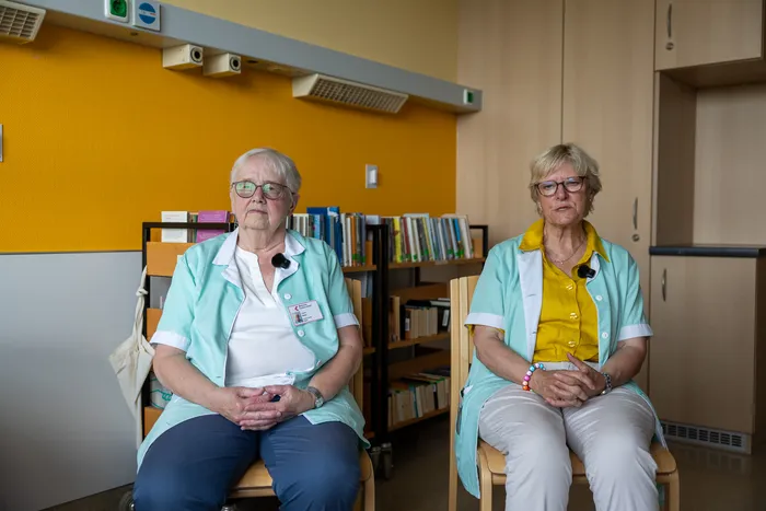 Zwei ältere Frauen in weißen Kitteln sitzen nebeneinander auf Stühlen. Im Hintergrund befindet sich ein Bücherregal in einem hellen Raum.