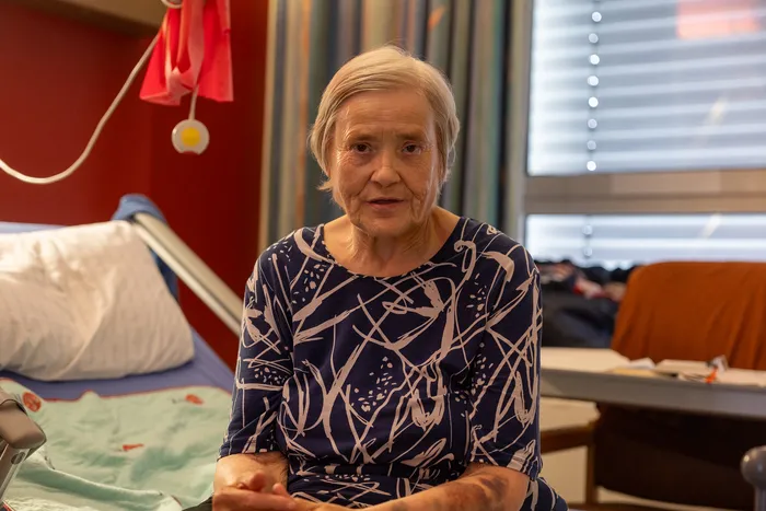 Eine ältere Frau mit weißem Haar blickt ernst in die Kamera. Sie sitzt in einem Krankenhausbett vor einem Fenster mit Jalousien.