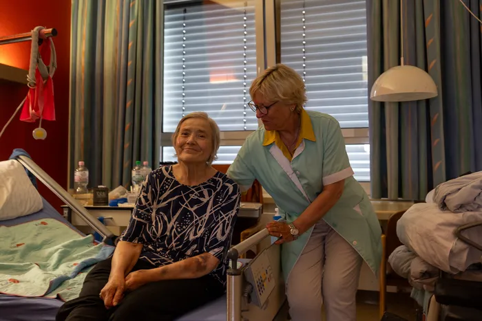 Eine ältere Patientin sitzt lächelnd in einem Krankenhausbett, während eine Frau in Uniform neben ihr steht und etwas auf ein Gerät sprüht. Im Hintergrund sind ein Fenster mit Vorhängen und ein weiteres Krankenhausbett zu sehen.