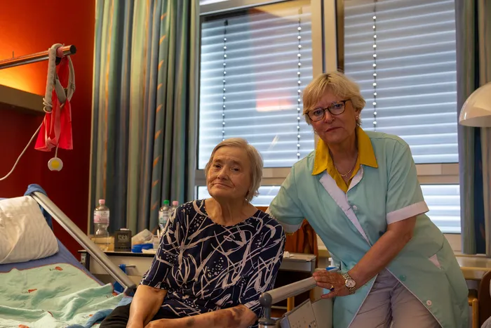 Eine ältere Frau sitzt im Krankenhausbett, neben ihr steht eine Frau in Uniform. Im Hintergrund sind Fenster mit Jalousien und ein roter Vorhang zu sehen.