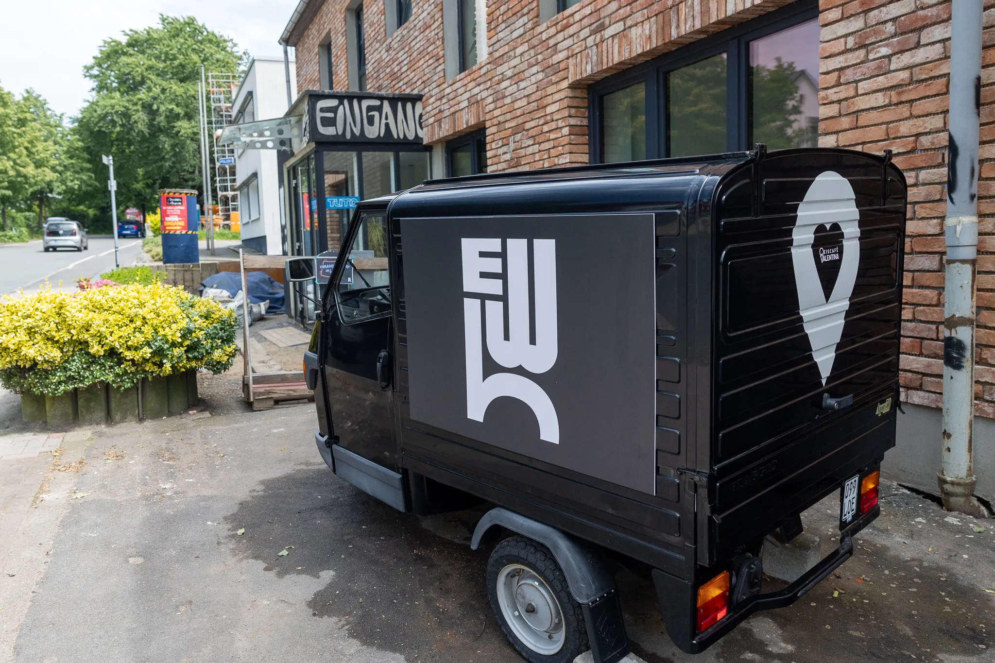 Schwarzer Transporter mit großem weißen „W“ und einem Standortsymbol mit Herz auf der Rückseite steht vor einem Gebäude mit dem Hinweis „Eingang“.
