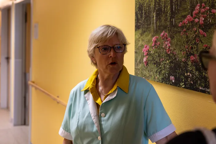 Eine Frau mit grauen Haaren und Brille in einem hellgrünen Arbeitskittel spricht in einem Krankenhausflur. Im Hintergrund ein großes Bild mit blühenden Rosen.