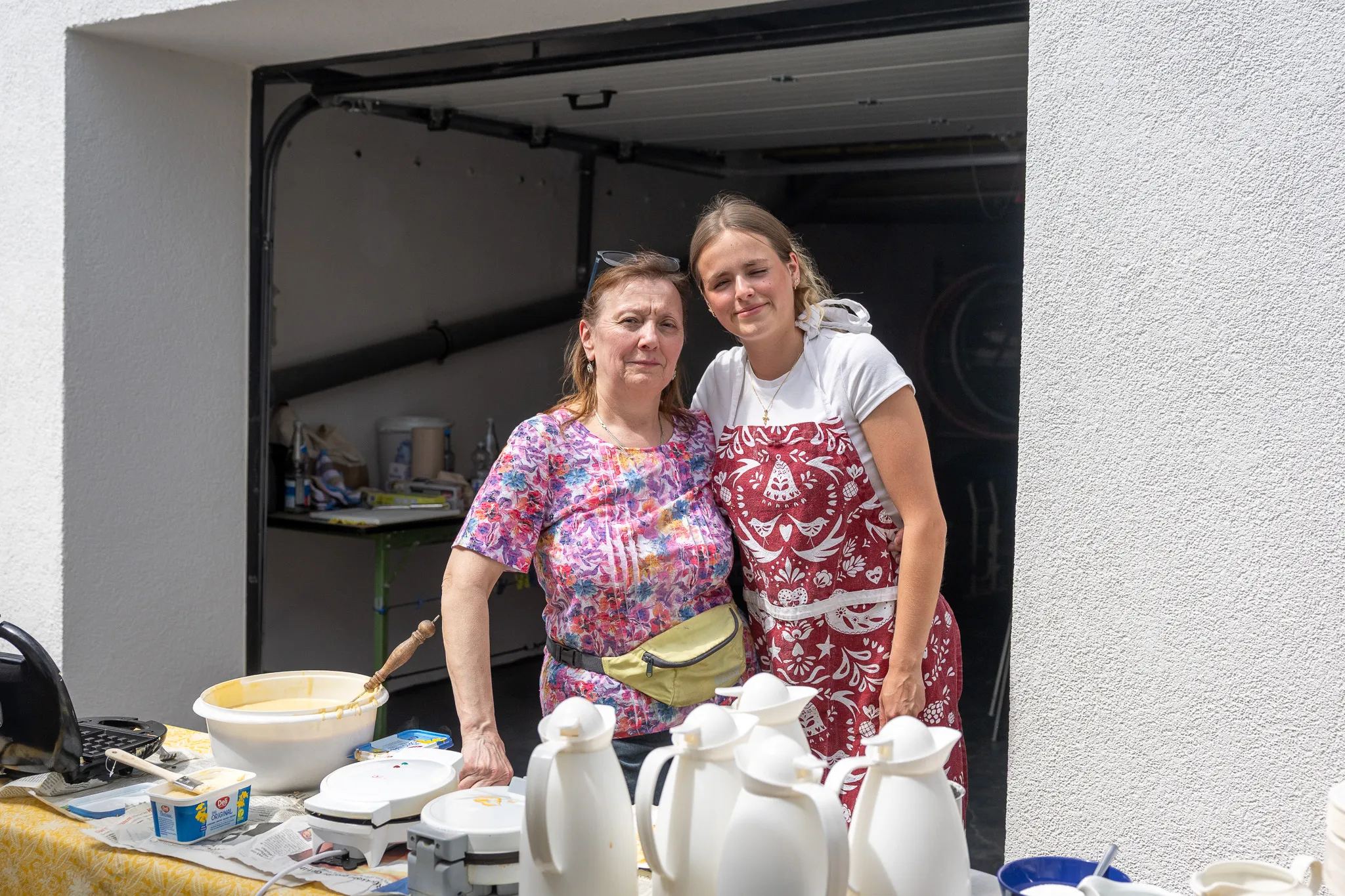 Zwei Frauen stehen lächelnd vor einem Tisch mit Gebäck und Kaffeekannen. Im Hintergrund ist eine offene Garage zu sehen.
