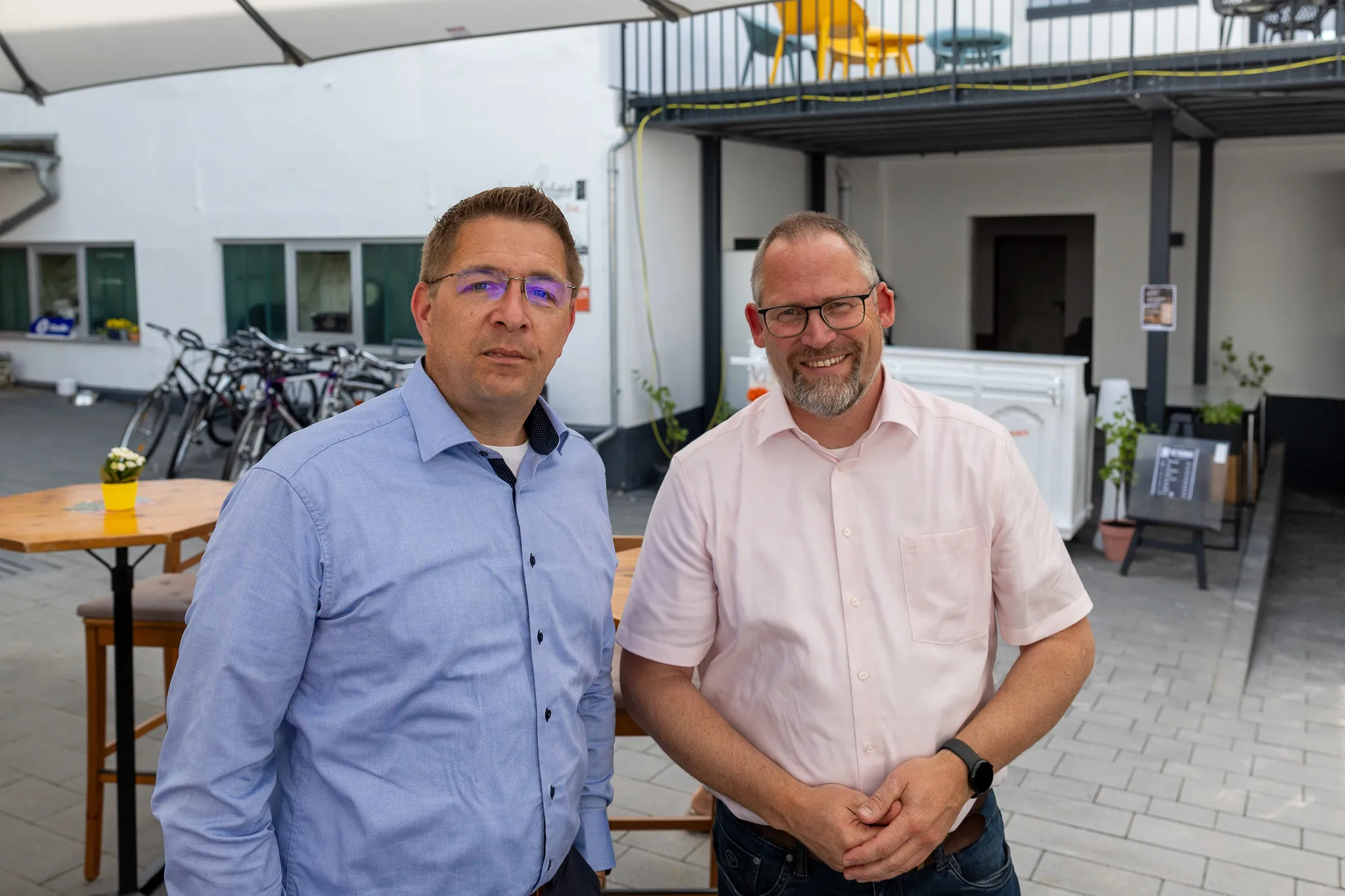 Zwei lächelnde Männer stehen vor einem Gebäude mit einer überdachten Terrasse. Im Hintergrund sind Fahrräder und gelbe Stühle zu sehen.