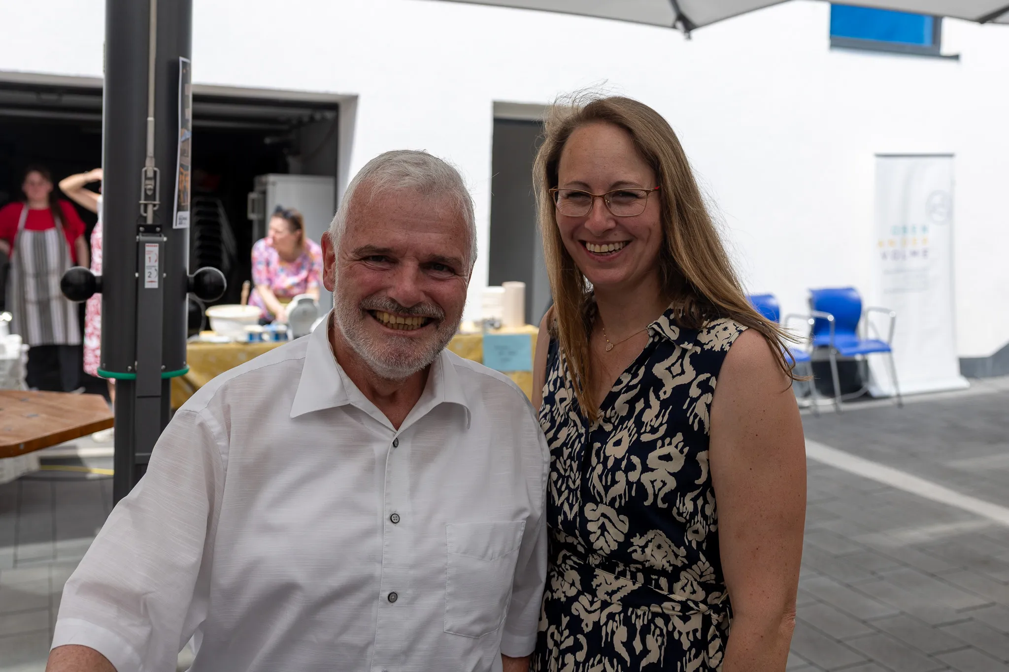 Zwei lächelnde Menschen, ein älterer Mann mit grauem Haar und eine jüngere Frau mit langen, blonden Haaren, stehen gemeinsam vor einer Markise. Im Hintergrund sind weitere Personen und ein Stand zu sehen.