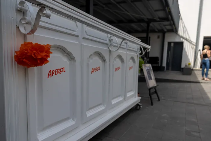 Weiße Bar mit dem Aufdruck „Aperol“ und einer orangefarbenen Dekoration, im Hintergrund Menschen und ein Werbeschild.