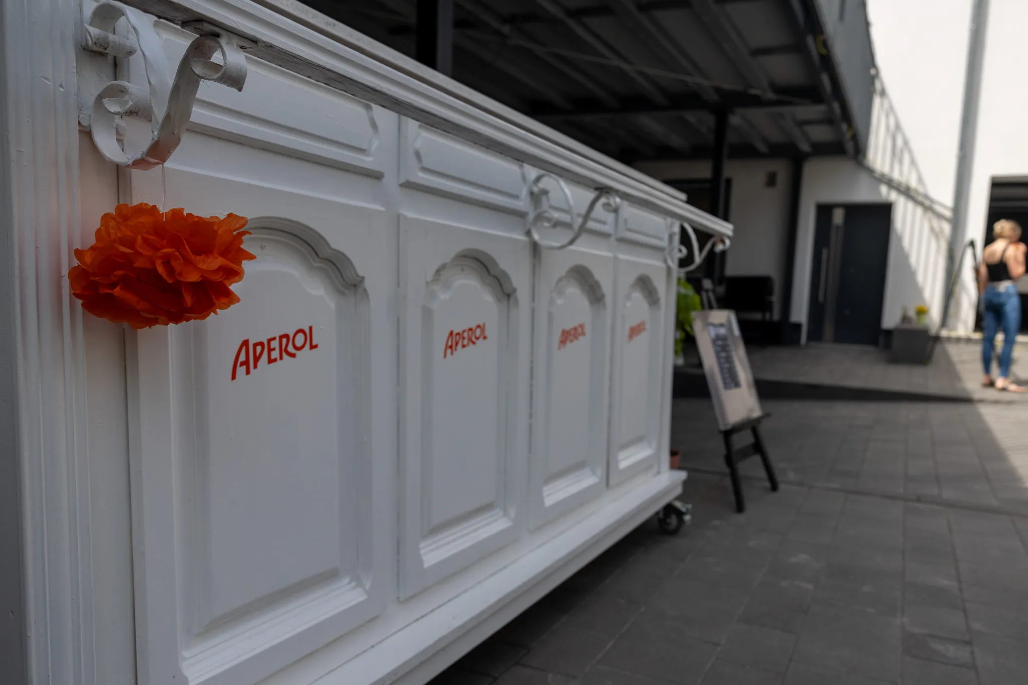 Weiße Bar mit dem Aufdruck „Aperol“ und einer orangefarbenen Dekoration, im Hintergrund Menschen und ein Werbeschild.