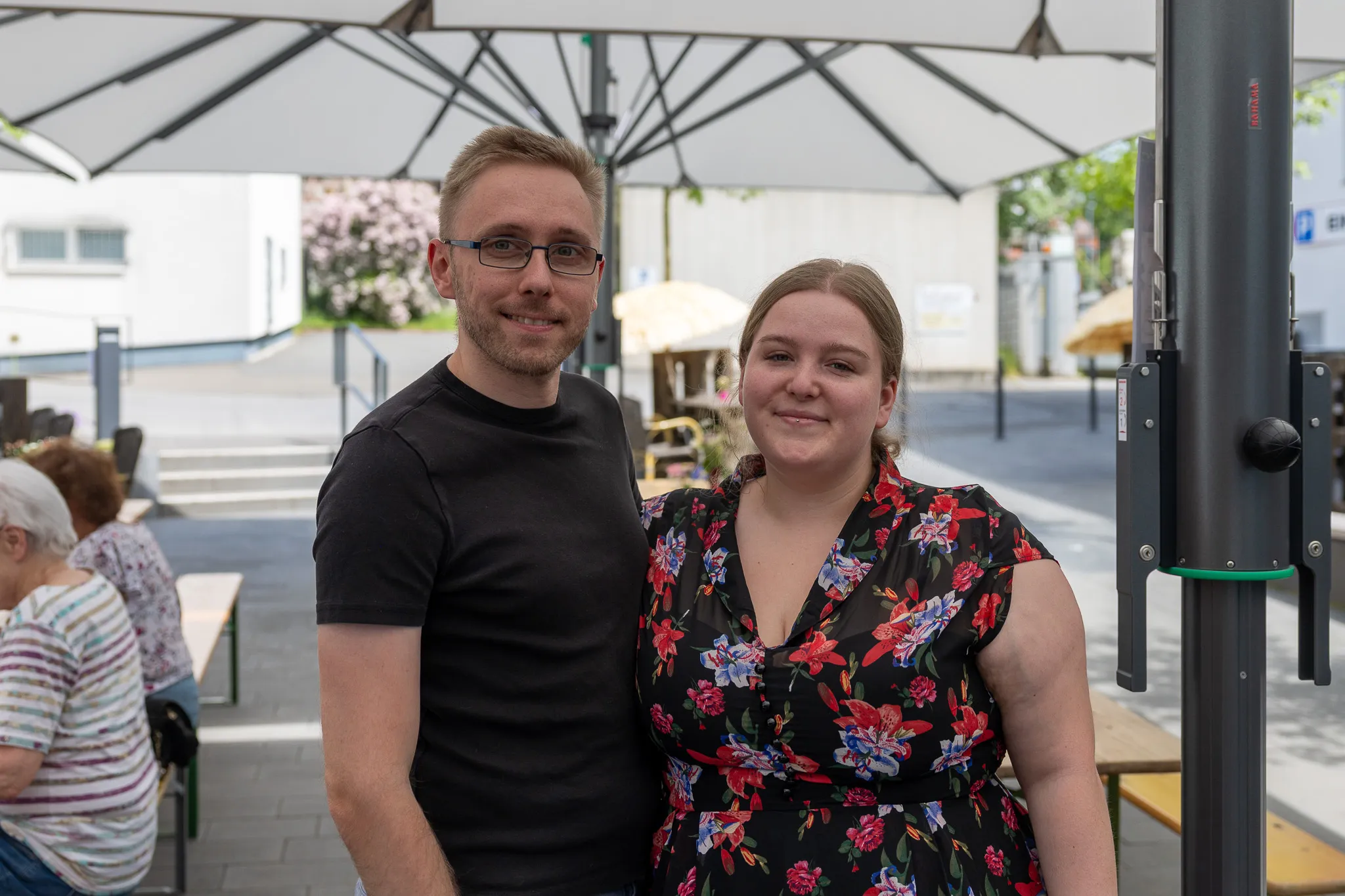 Ein Mann und eine Frau lächeln in die Kamera, stehen vor einer grauen Säule und Sonnenschirmen im Freien.