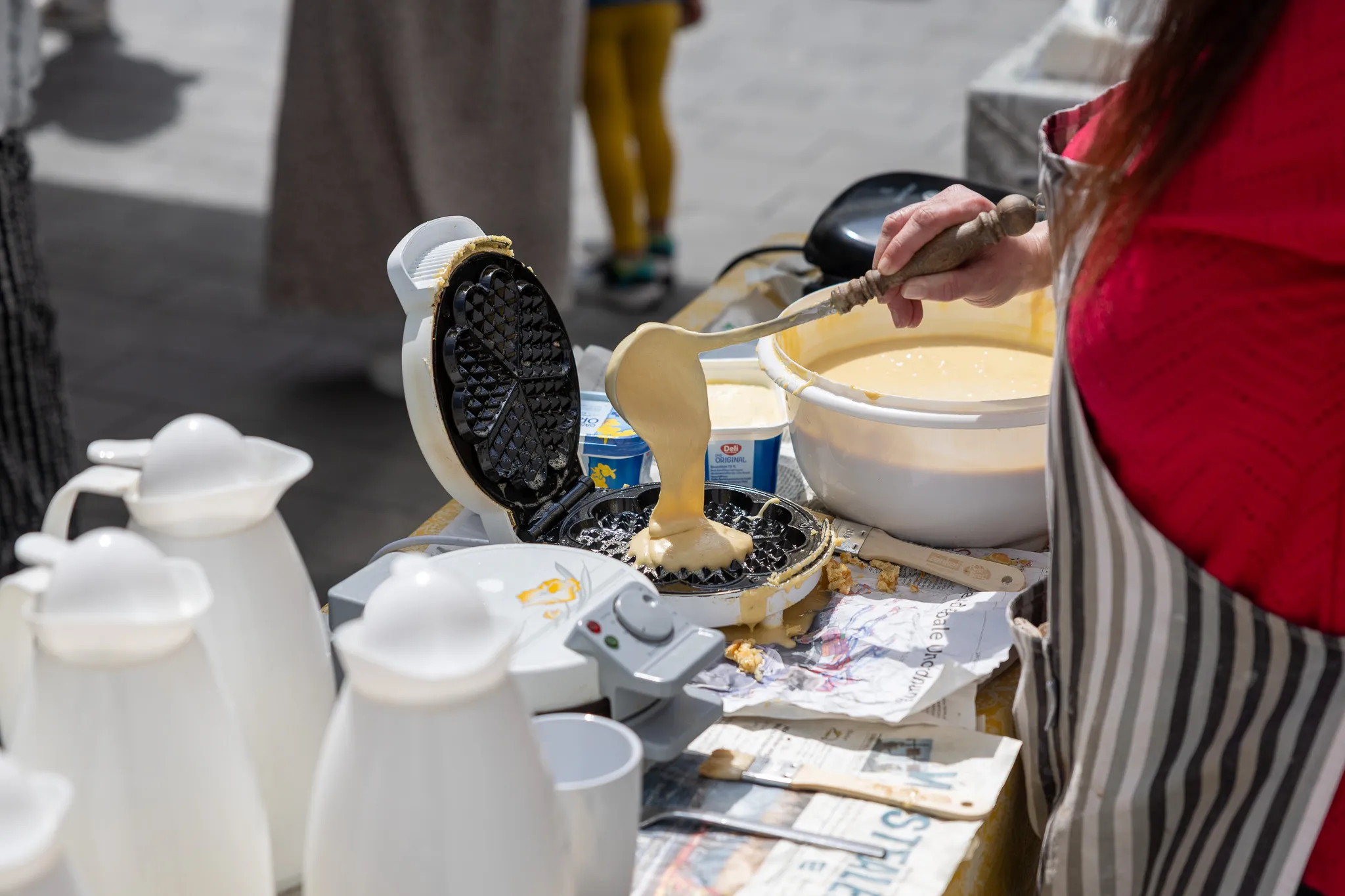 Eine Person gießt Teig aus einer Schüssel auf ein Waffeleisen, während daneben Becher und Butter bereitstehen.