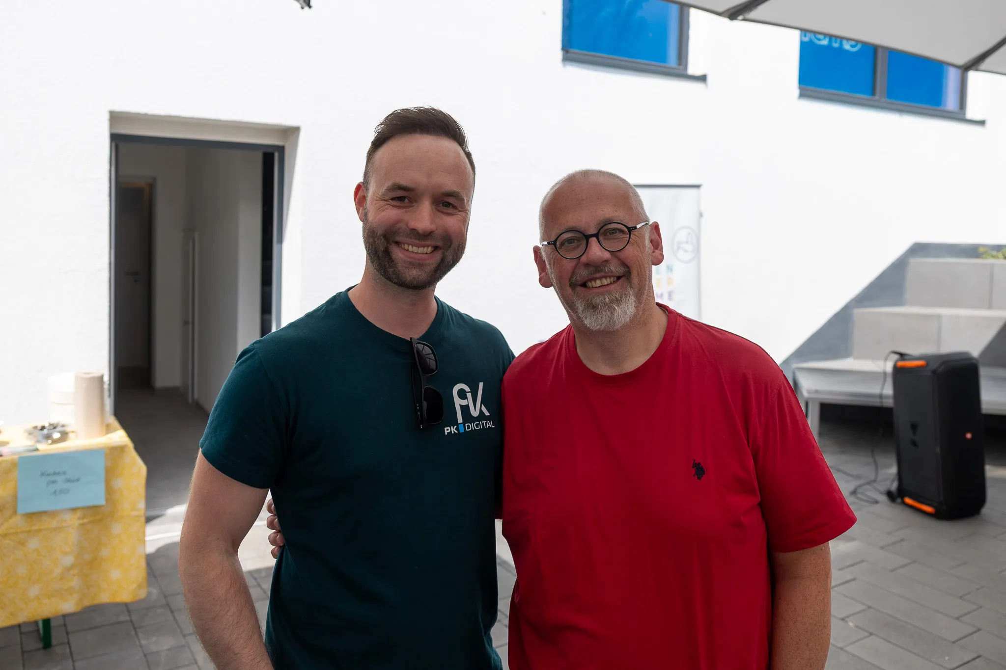 Zwei lächelnde Männer posieren vor einer hellen Fassade. Der Mann links trägt ein dunkelgrünes Shirt mit dem Logo „PK DIGITAL“, der Mann rechts ein rotes Poloshirt.
