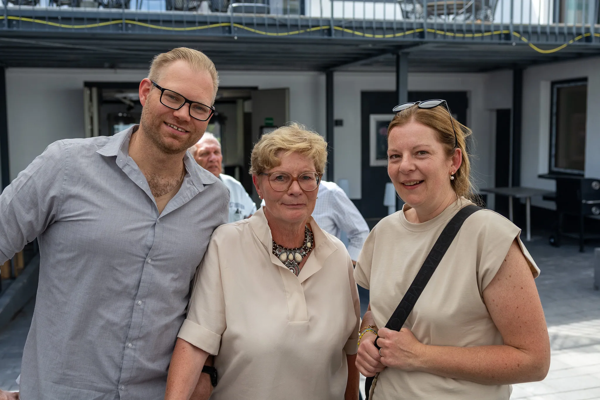 Drei Personen lächeln in die Kamera: ein Mann mit Bart und Brille, eine ältere Frau mit Perlenkette und eine jüngere Frau mit hochgestecktem Haar. Im Hintergrund ein Innenhof mit weiteren Menschen.