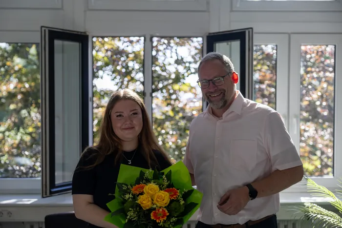 Jolina Klotz hält einen Blumenstrauß vor einem Mann mit Brille und Hemd. Im Hintergrund sind Fenster und Bäume zu sehen.
