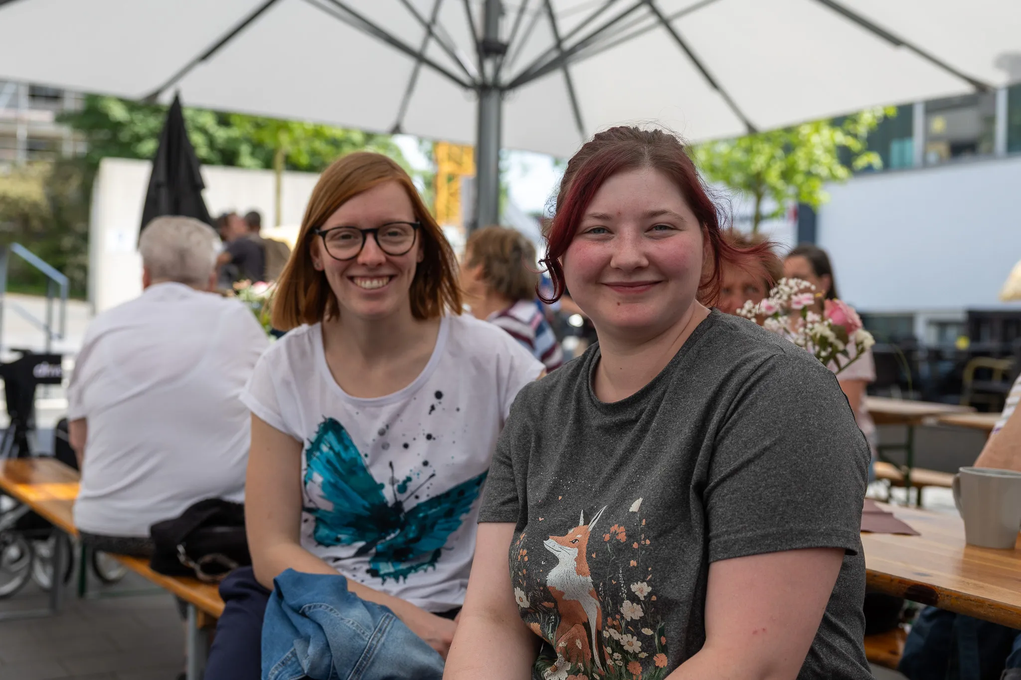 Zwei junge Frauen lächeln in die Kamera, sitzen an einem Tisch im Freien unter einem Sonnenschirm. Im Hintergrund sind weitere Menschen und ein Gebäude zu sehen.
