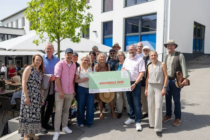 Gruppe von etwa zwanzig Menschen steht lächelnd vor einem modernen Gebäude mit dem Banner „Südwestfalen REGIONALE 2025“.