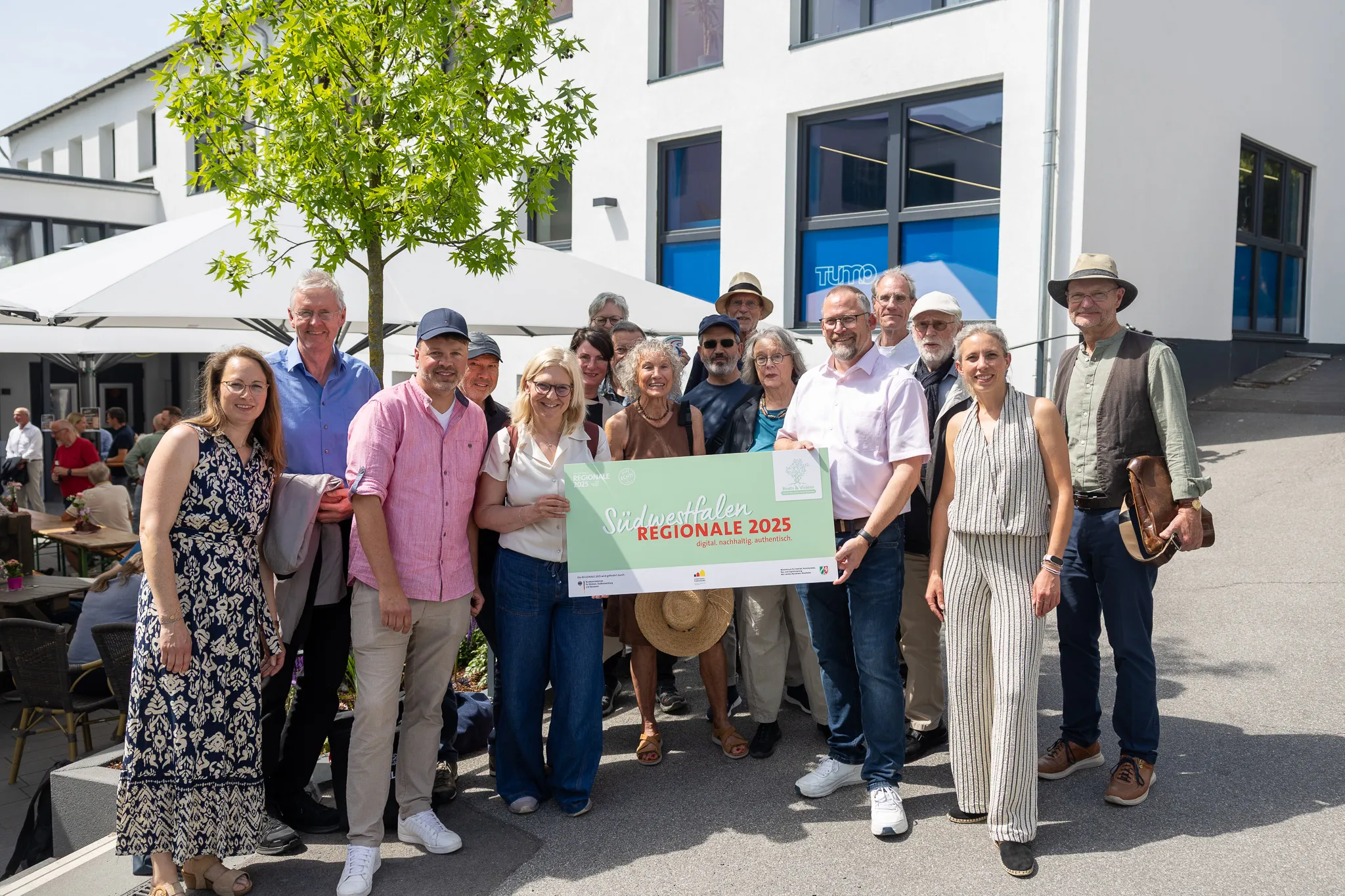 Gruppe von etwa zwanzig Menschen steht lächelnd vor einem modernen Gebäude mit dem Banner „Südwestfalen REGIONALE 2025“.