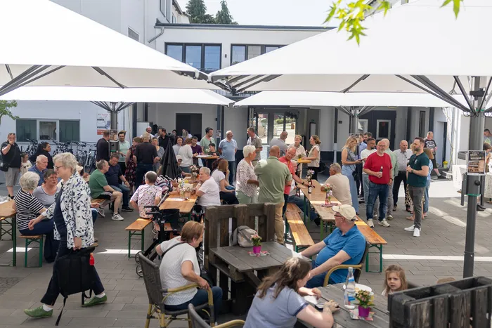 Menschengruppen sitzen und stehen an Biertischgarnituren vor einem hellen Gebäude mit weißen Sonnenschirmen. Im Hintergrund sind weitere Personen und ein Eingangsbereich zu sehen.