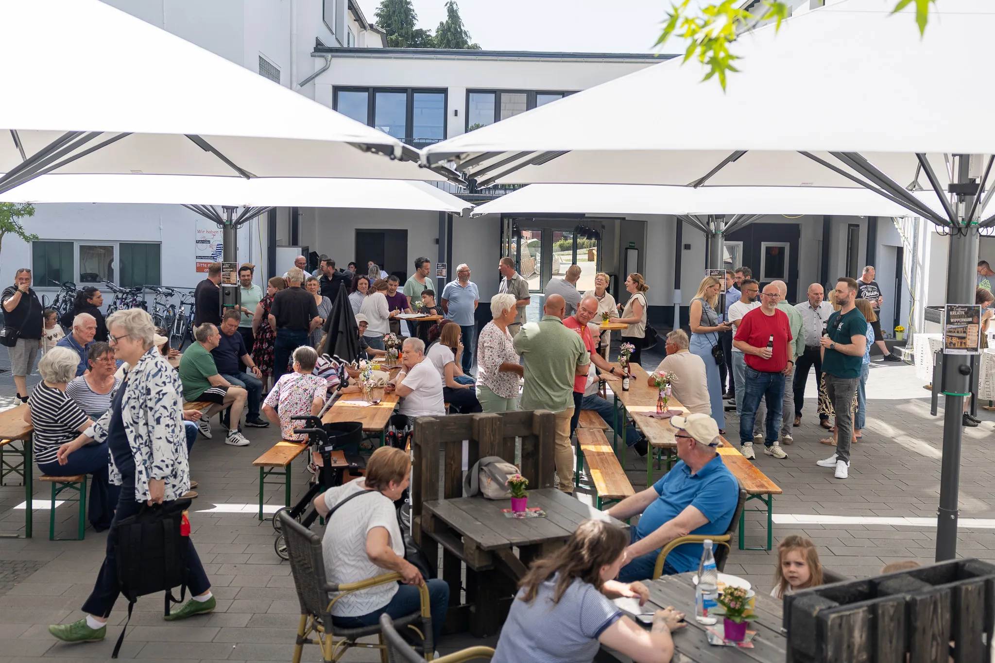 Menschengruppen sitzen und stehen an Biertischgarnituren vor einem hellen Gebäude mit weißen Sonnenschirmen. Im Hintergrund sind weitere Personen und ein Eingangsbereich zu sehen.